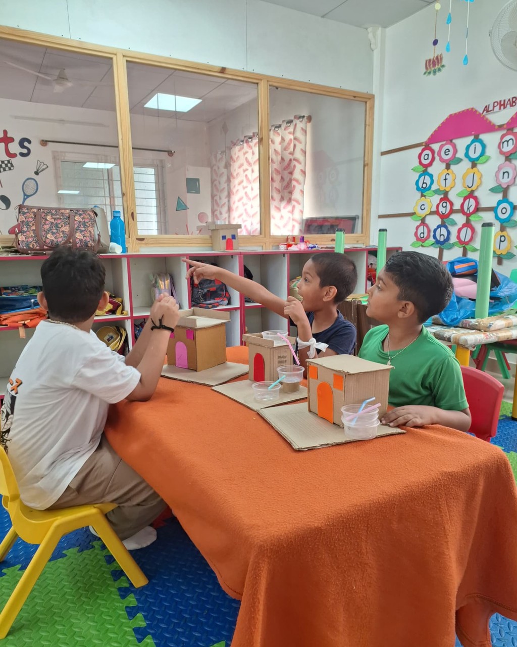 Three children collaborating around a classroom table with cardboard house models and craft materials